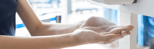 Person hält Hände unter einem Spender, um Flüssigkeit aufzunehmen, in heller Umgebung nahe einer Wand montiert