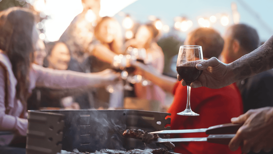 Fleisch wird gegrillt mit einem Glas Wein in der Hand und im Hintergrund stoßen Menschen mit ihren Gläsern an