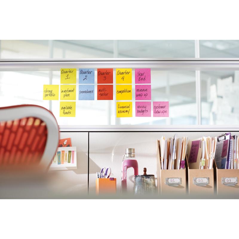 Egy sor 3M Post-it® jegyzetlap, sárga, 76x76 mm (90 lap/blokk, 6 blokk/csomag) egy üveg elválasztón van elrendezve; alatta irodai asztalon mappák, könyvek, egy vizespalack és más eszközök találhatók.