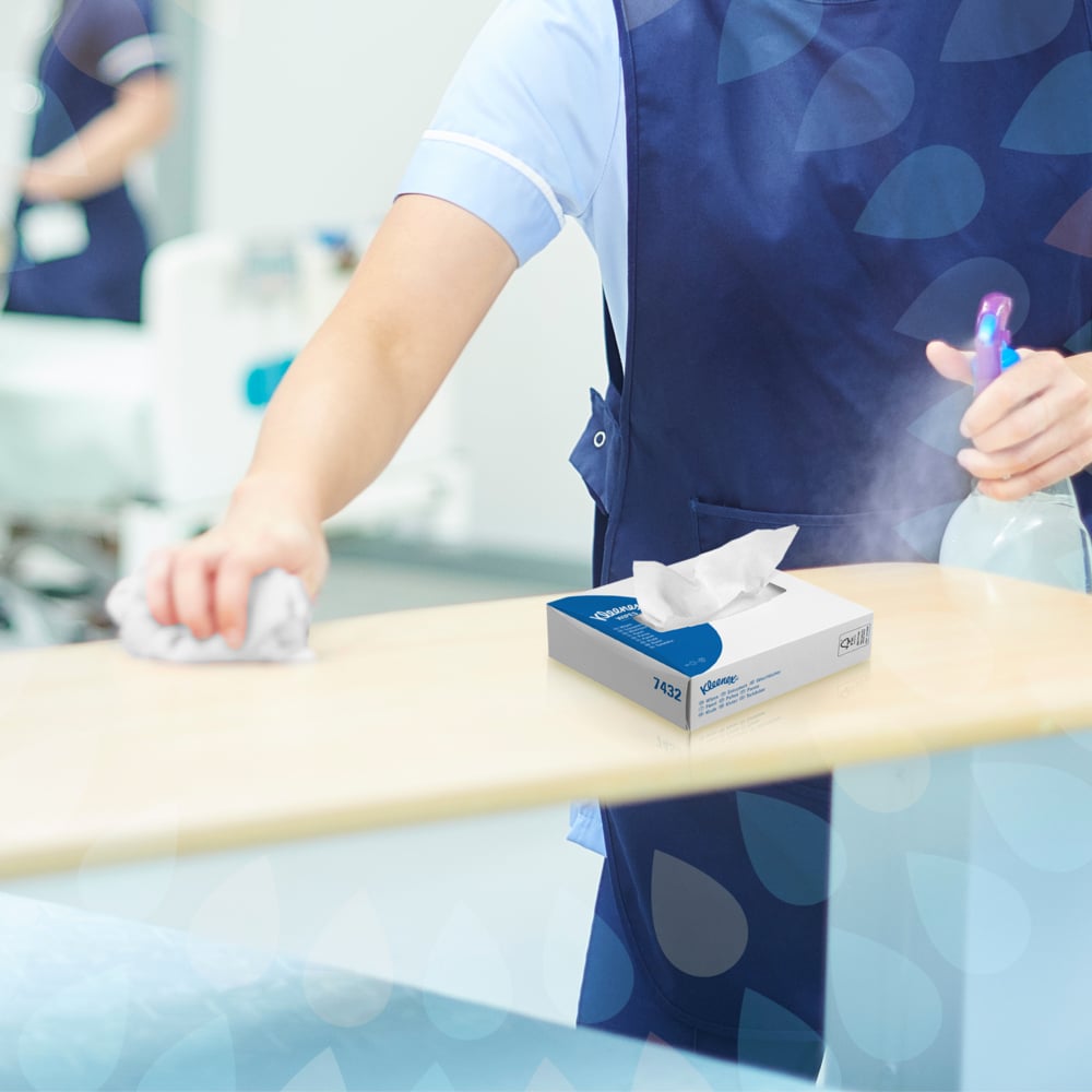 Eine Person in blauer Uniform wischt einen Tisch mit einem Tuch ab und hält eine Sprühflasche in der Hand. Auf dem Tisch steht eine Schachtel Kleenex® Wischtücher Interfold (66 x 80) der Kimberly-Clark GmbH, die auf eine hygienische Reinigung in dem hellen Raum hinweist.