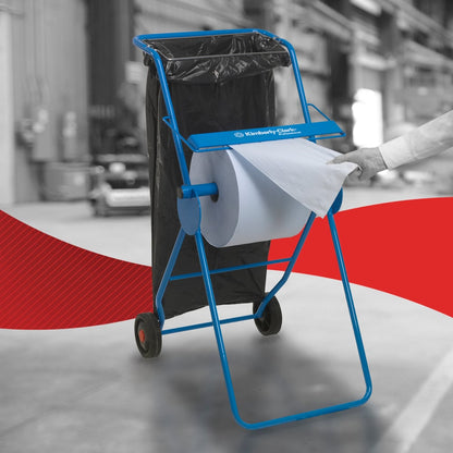 Egy személy egy WypAll® L10 papírtörlőt húz felületekhez - nagy tekercs (38,00 x 23,50 cm) a Kimberly-Clark GmbH-tól egy kék fémállványról kerekekkel és egy hozzáerősített fekete szemeteszsákkal, ami hatékony felülettisztítást jelez ipari környezetekben.