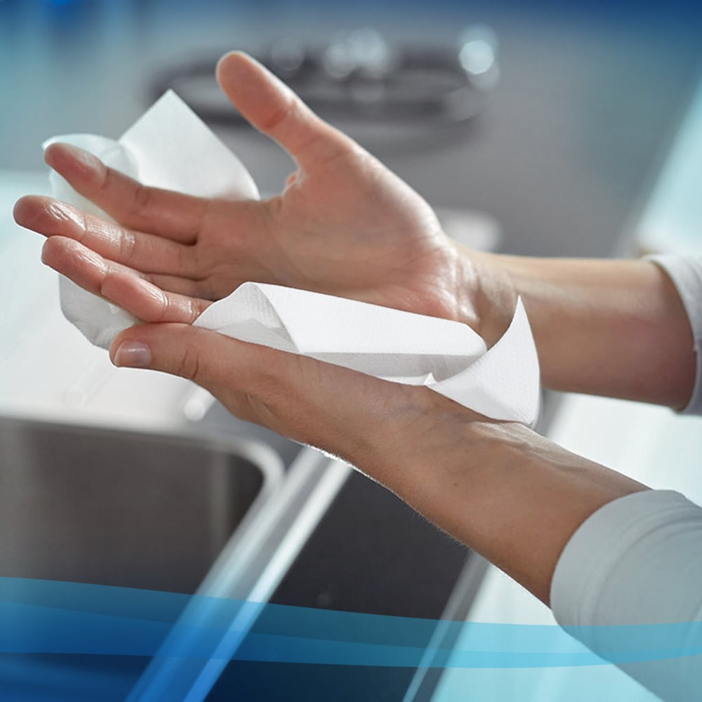 Egy személy a kezét szárítja egy mosogató felett Scott® kéztörlőkkel - MultiFold / Fehér (16 csomag x 250 lap | karton) a Kimberly-Clark GmbH-tól, elmosódott munkalapokkal és konyhai tárgyakkal a háttérben.