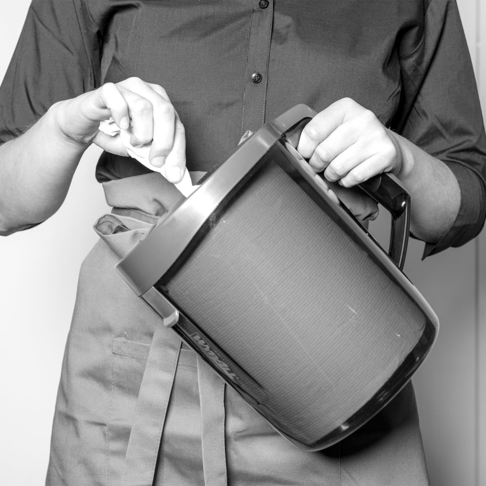 Egy kötényes és begombolt inges személy egy nagy kancsót kever, miközben egy WypAll® Reach™ Centrefeed adagoló (fekete) a Kimberly-Clark GmbH-tól a közelben áll, professzionális megjelenést kölcsönözve. A fekete-fehér kép kiemeli a jelenetet.