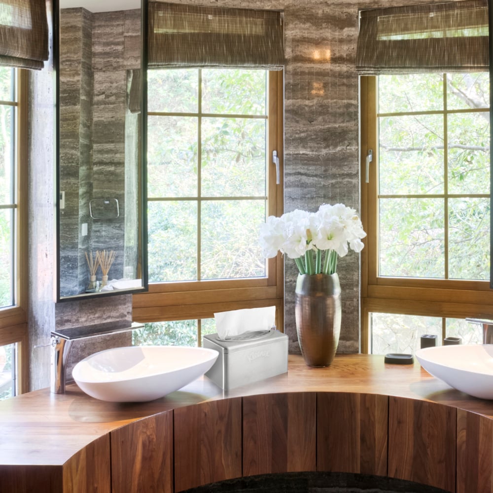 Egy modern fürdőszoba fa munkalappal, két fehér mosdóval, barna redőnyökkel, egy vázával fehér virágokkal és egy elegáns Kimberly-Clark GmbH rozsdamentes acélból készült Kleenex® kéztörlő adagolóval kihúzható doboz funkcióval a zsebkendődoboz mellett. Kint fák láthatók.