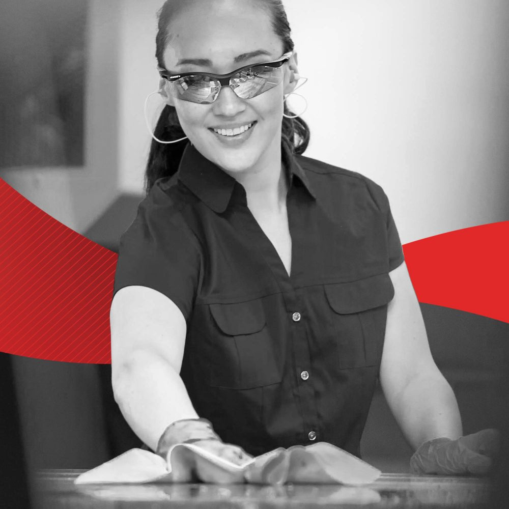 Egy kesztyűt és szemüveget viselő nő mosolyog, miközben egy felületet tisztít egy WypAll® X60 General Clean - BRAG™ Box kendővel (41,80 × 28,30 cm) a Kimberly-Clark GmbH-tól; a háttérben egy piros grafikai dizájn jelenik meg.