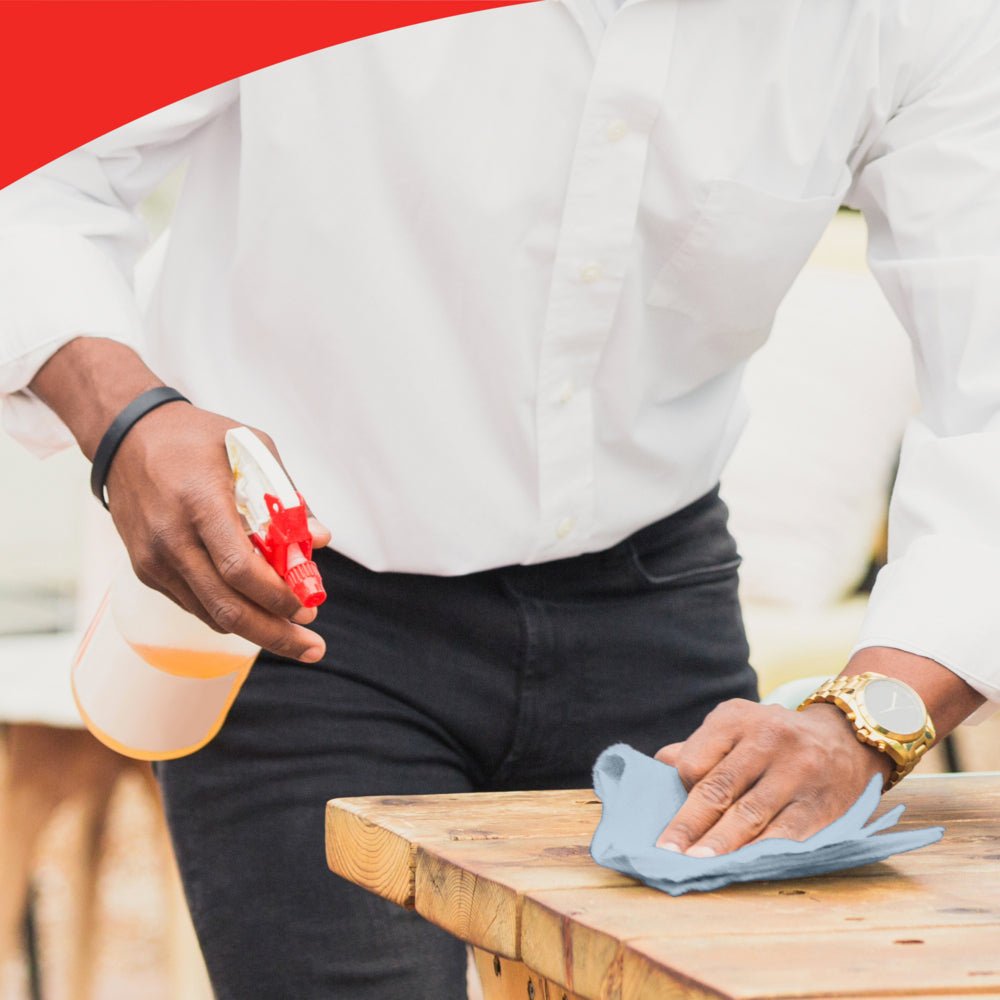 Egy személy fehér ingben és órával tisztít egy faasztalt egy kék kendővel, valószínűleg WypAll® L20 General Clean - BRAG™ Box a Kimberly-Clark GmbH-tól. A kép tetején egy piros, ívelt szegély található, amely a részletekre való figyelmet hangsúlyozza.