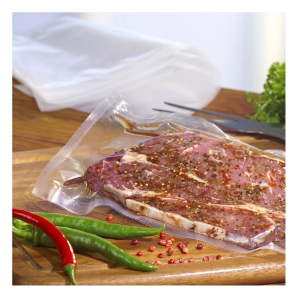 Ein rohes, mariniertes Steak wird in einem transparenten Starpak 100 Siegelrandbeutel (PA/PE) mit einem Kammervakuumierer auf einem Holzbrett vakuumiert, mit Chilischoten, rosa Pfefferkörnern, Blattgemüse und einer Gabel im Hintergrund.