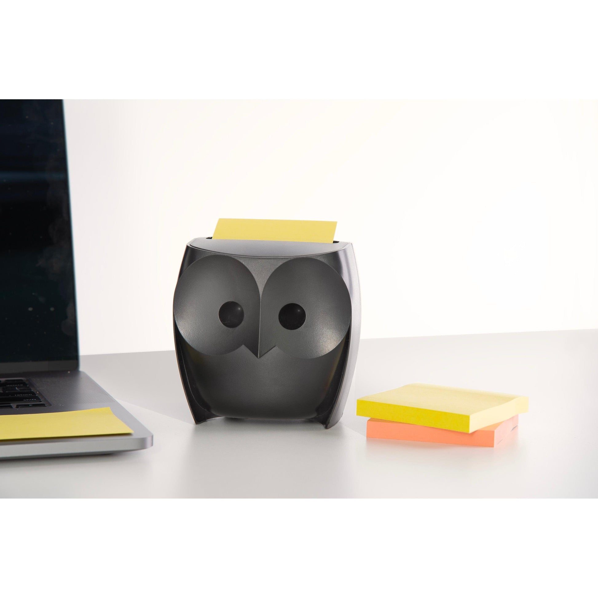 Ein schwarzer, eulenförmiger Post-it® Z-Notes Dispenser der 3M Deutschland GmbH mit einer gelben Notiz liegt auf einem Schreibtisch neben zwei Packungen Post-it® Super Sticky Z-Notes (45 Blatt/Block, 2 Blöcke/Packung), neben einem aufgeklappten Laptop.