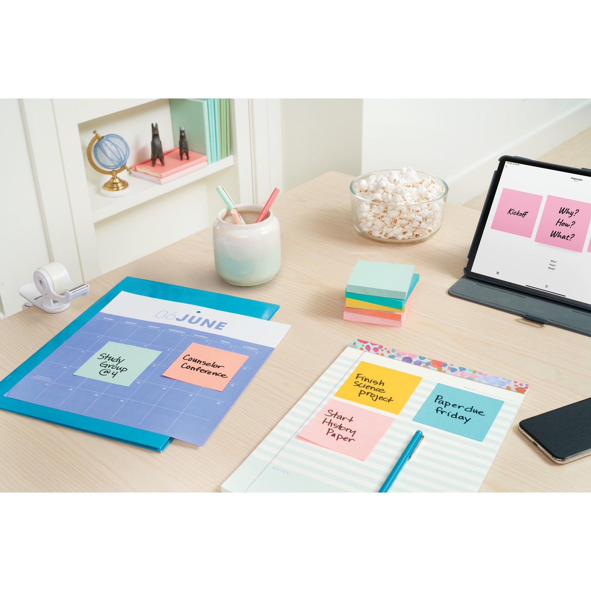 Ein Schreibtisch mit Post-it® Notes Beachside Collection von 3M, einem Juni-Kalender, Erinnerungen, einem Notizbuch, einem Popcornglas, einem iPad mit Notizen, Klebeband, Stiften in einer Tasse und Regalen mit Bürobedarf im Hintergrund.