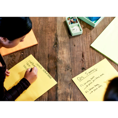 Két személy ül egy faasztalnál és írnak a 3M Post-it® Super Sticky Big Notes BN11-EU, Sárga (279 x 279 mm, 30 lap/blokk, 100% PEFC) jegyzetlapokra, és ötleteket agyalnak ki, mint például "FOTÓZÁS CIPŐ STRATÉGIA" és "Q4 Stratégia". Sárga jegyzetlapok és markerek állnak készen.