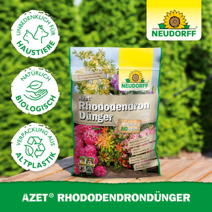 Ein Beutel Azet RhododendronDünger aus dem Neudorff Shop liegt auf einer Holzfläche im Freien vor grünen Hecken. Auf der Verpackung aus Biokunststoff steht, dass er biologisch, haustierfreundlich und ideal für Rhododendren und Azaleen ist.