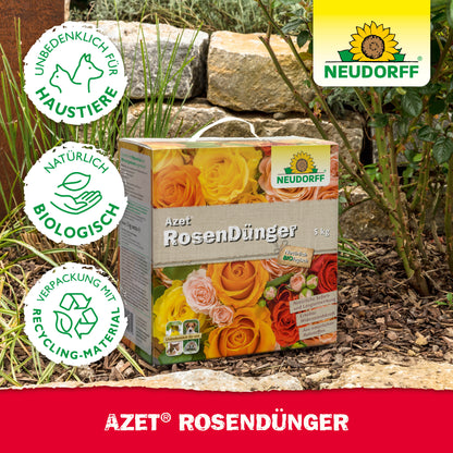 Eine Schachtel Neudorff Shop Azet RosenDünger liegt in einem Garten zwischen Steinen und Pflanzen. Die Verpackung betont die Sicherheit von Haustieren, natürliche Inhaltsstoffe und recycelbare Materialien für blühende Rosen.
