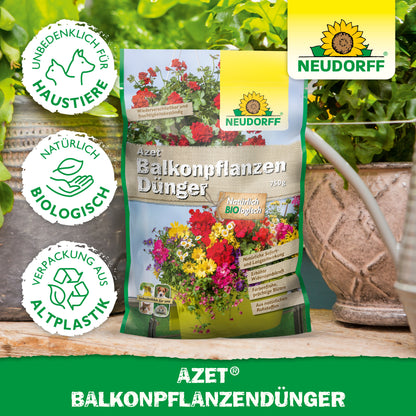 Eine Tüte Azet BalkonpflanzenDünger von Neudorff Shop steht auf einem Tisch zwischen Topfpflanzen und Balkonkästen. Die Verpackung zeigt Blumen, Text und Symbole für Tierschutz, biologische Inhaltsstoffe und Verpackungen aus Biokunststoff.