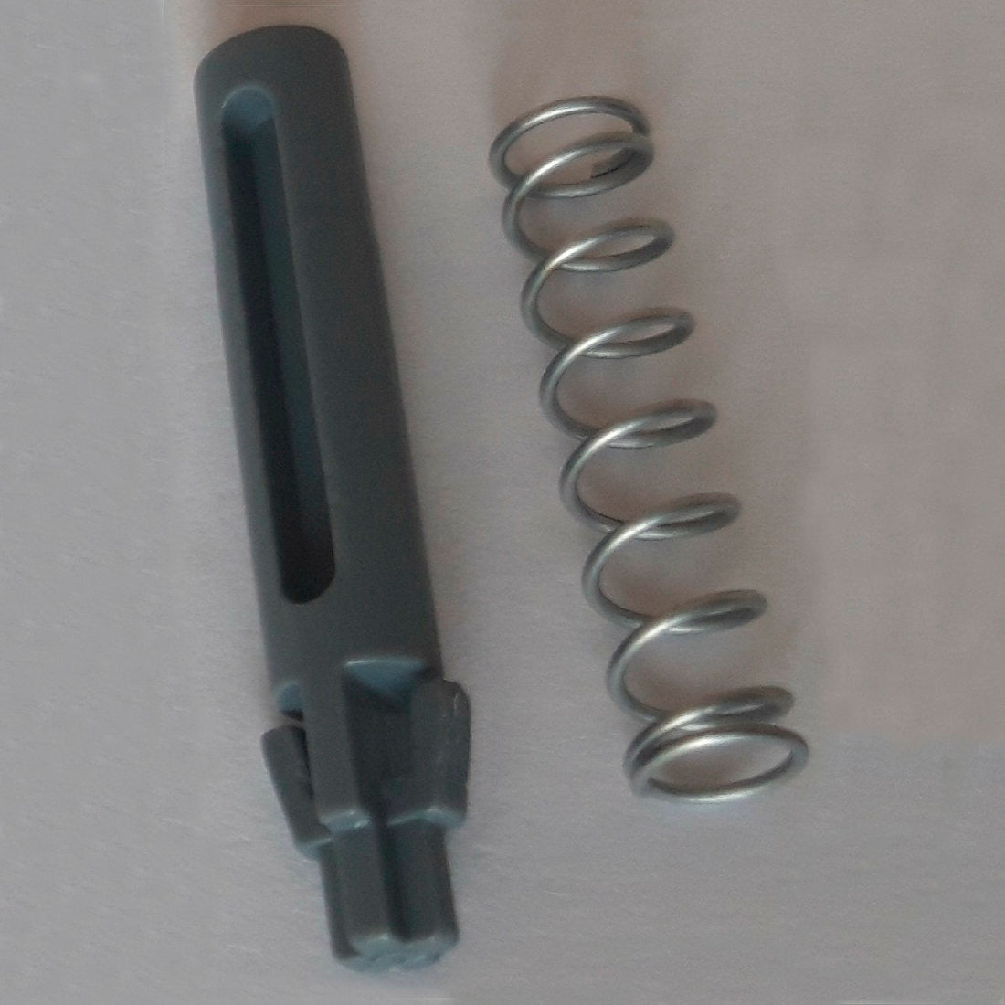 Egy fémrugó és egy sötétszürke hengeres pótkilincs, amely barázdákkal és fogakkal van ellátva, a Diversey Deutschland GmbH & Co. OHG DI Protect kilincs & rugó (1 darab) részét képezi, világos felületen van ábrázolva, és bemutatja a DI Protect kocsi robusztus anyagait.