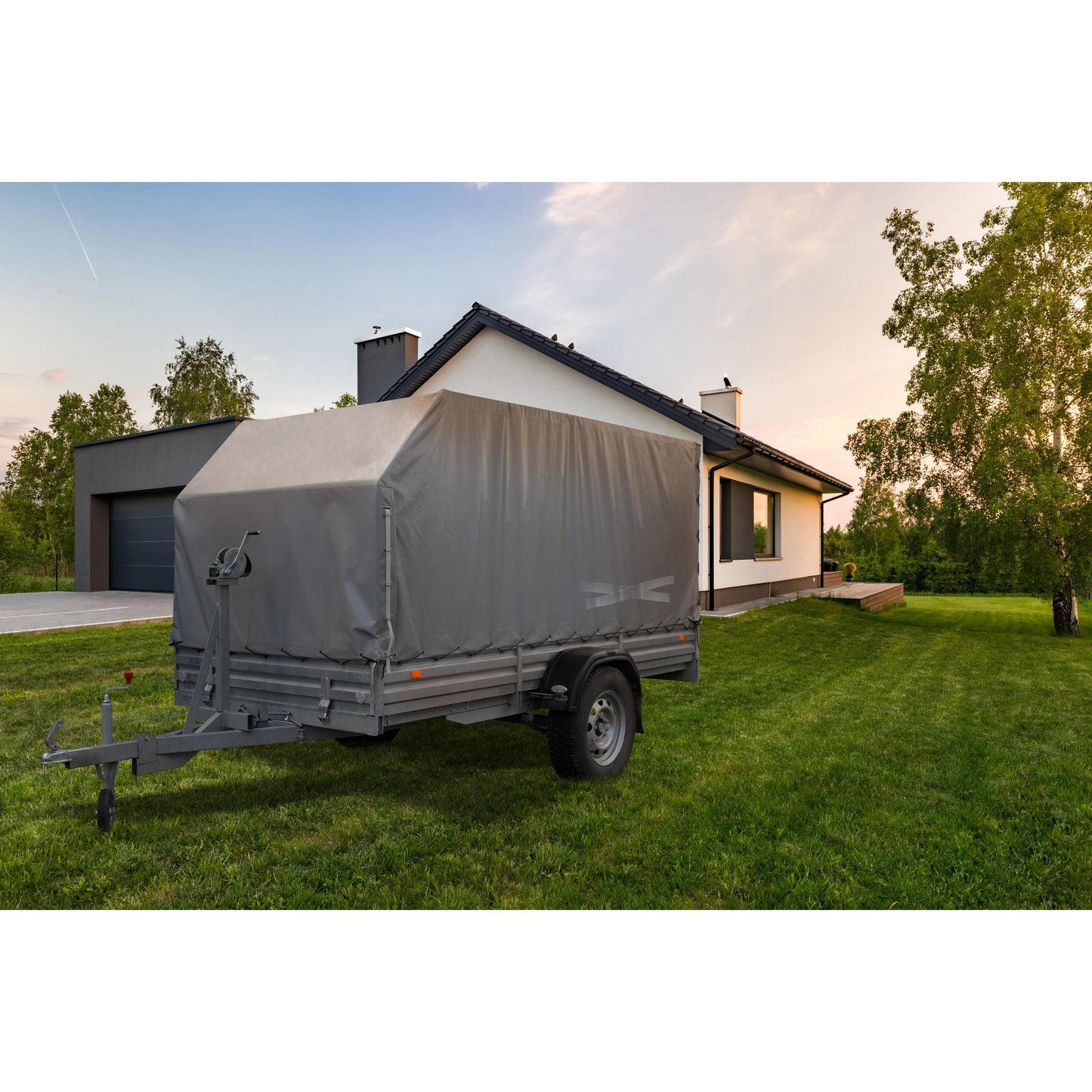 Ein grauer Nutzfahrzeuganhänger, der mit dem Scotch® Extremium™ All Weather Hochleistungs-Klebeband (27m x 48mm) der 3M Deutschland GmbH gesichert ist, steht bei teilweise bewölktem Himmel auf einer Wiese vor einem modernen weißen Haus mit dunklem Dach und angeschlossener Garage.