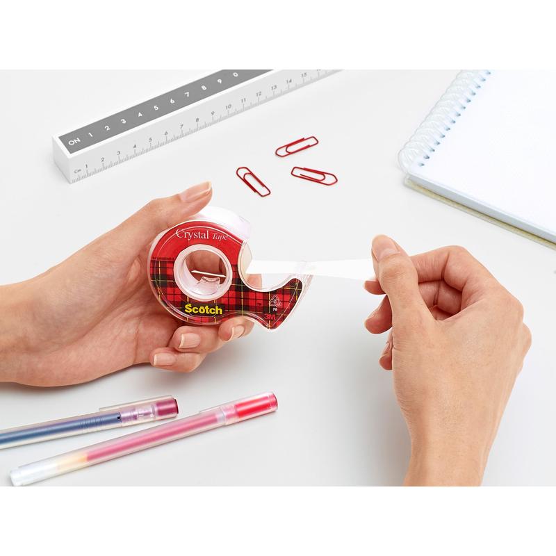 Egy személy a 3M Deutschland GmbH piros kézi adagolójából húzza ki a Scotch® Crystal ragasztószalagot (19 mm x 7,5 m) egy fehér íróasztalon, piros gemkapcsokkal, tollakkal, egy vonalzóval és egy spirálfüzettel.