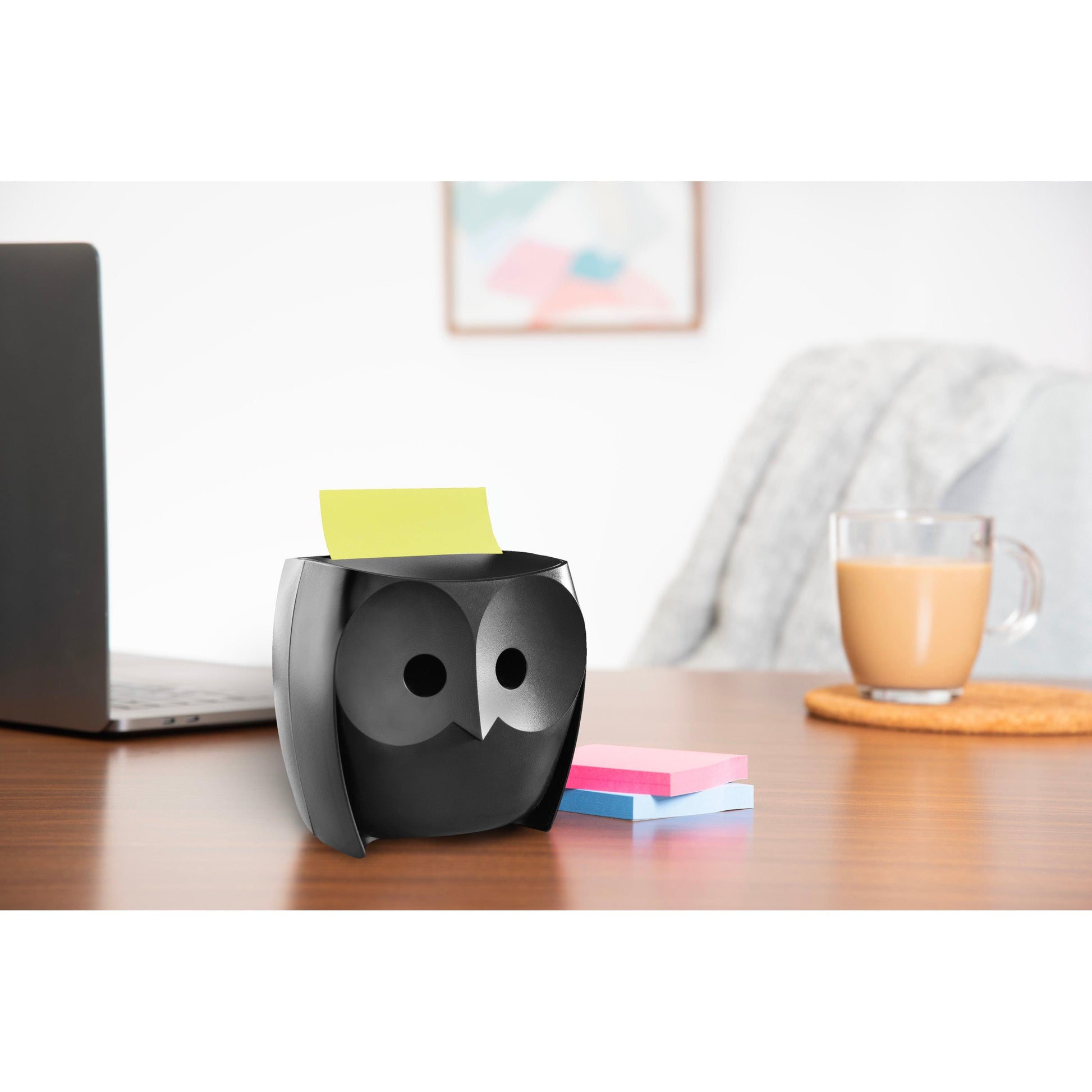 Auf dem Schreibtisch steht ein 3M Post-it® Z-Notes Dispenser Eule, Schwarz, mit gelben Super Sticky Z-Notes (45 Blatt/Block, 2 Blöcke/Packung), daneben rosa und blaue Zettel. Im Hintergrund sind ein Laptop, eine Tasse und eine graue Decke zu sehen.