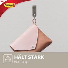 Ein mittelgroßer Command™ Metallhaken in gebürstetem Nickel von 3M hält eine rosa-braune Tasche (bis 1,3 kg) an einer hellgrauen Wand. Das Command-Logo ist sichtbar; der Haken wird ohne Bohren befestigt.