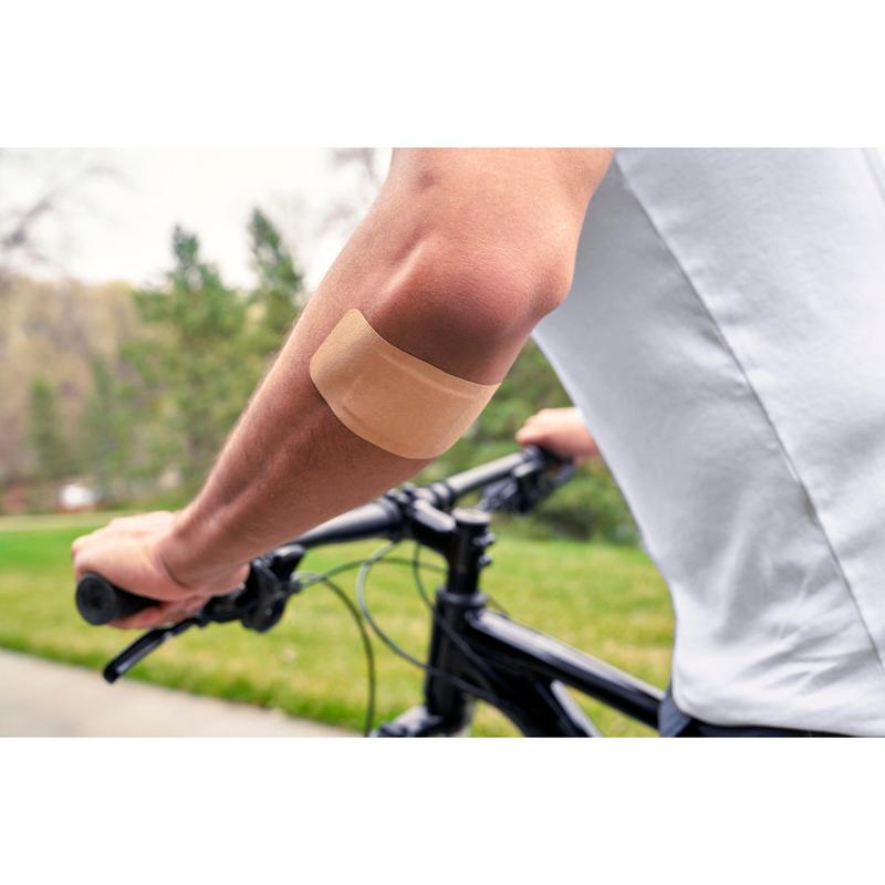 Egy személy, aki fehér inget visel és a szabadban biciklizik, egy Nexcare™ Ultra Stretch MAXI Comfort Flexible sebtapaszt (50 mm x 101 mm) a 3M Deutschland GmbH-tól a meztelen felkarján visel; a háttérben zöld fű és fák láthatók.