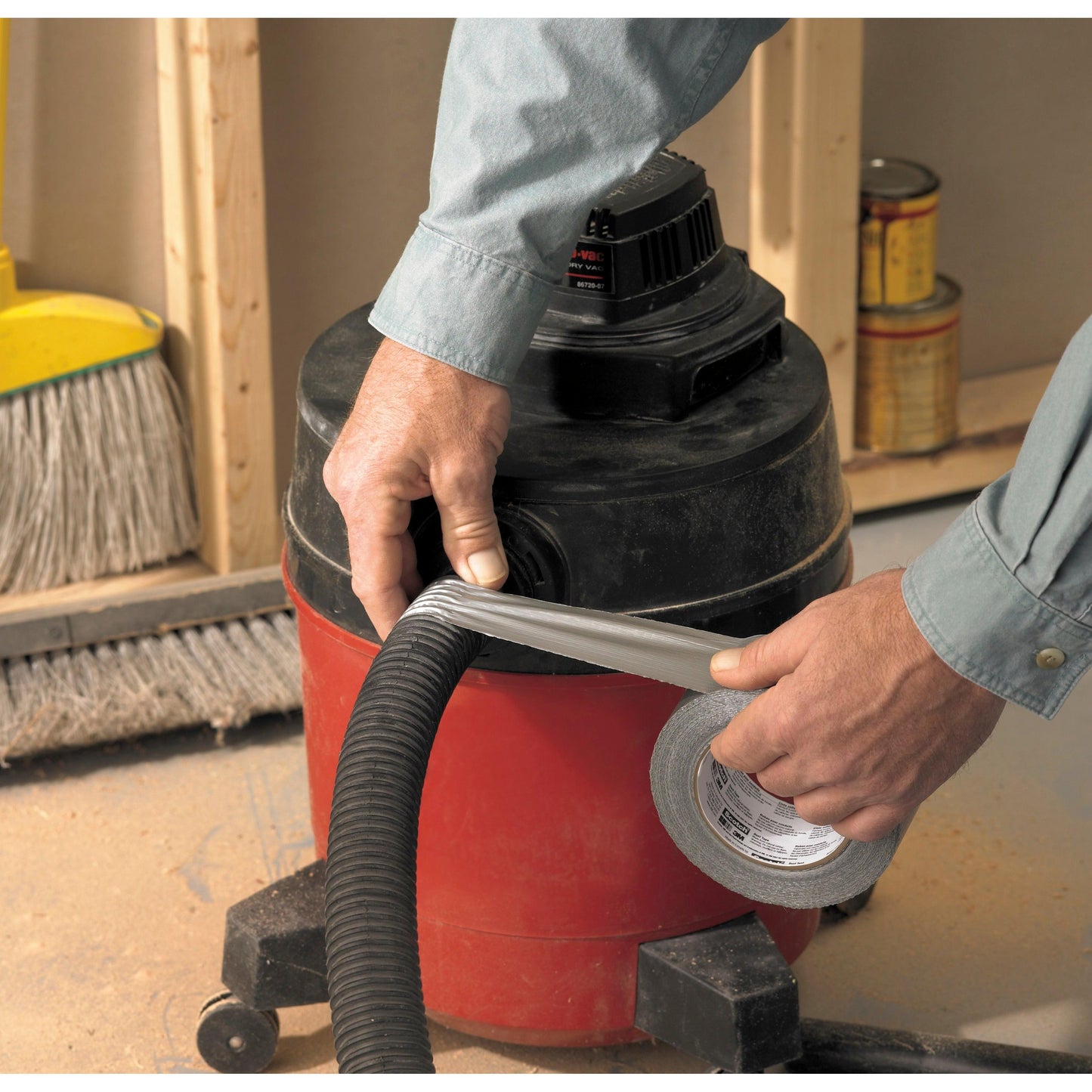 Egy személy Scotch® Universal ragasztószalagot 2904 (50 m x 48 mm) a 3M Deutschland GmbH-tól teker egy porszívócső köré, amely egy piros műhelyporszívóhoz van csatlakoztatva. A háttérben egy seprű és festékes dobozok láthatók.