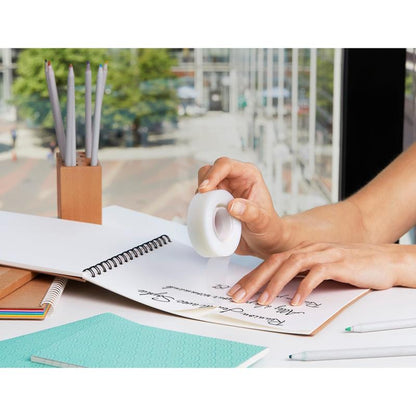 Egy személy javítószalagot használ kézzel írt jegyzetek felett egy spirálfüzetben, körülötte irodai kellékek, mint ceruzák, golyóstollak és a 3M Deutschland GmbH Scotch® Magic™ ragasztószalag (66% növényi alapú ragasztó, 19 mm x 33 m) egy ablak mellett, kilátással egy városi utcára.