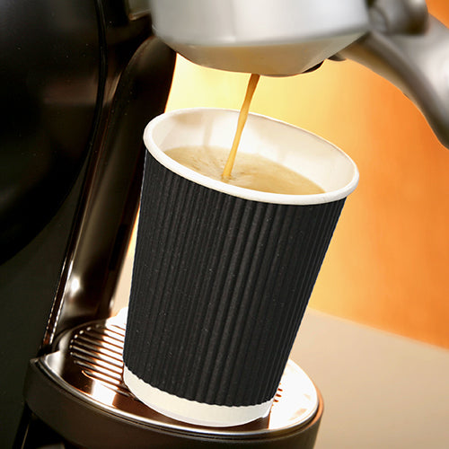 Ein Starpak 25 Trinkbecher, Pappe „To Go“ Ripple Wall-Becher steht auf einer Metalltropfschale unter dem Auslauf einer Kaffeemaschine, während heißer Kaffee hineinfließt, und hebt sich vor einem sanft beleuchteten, warm getönten Hintergrund ab.