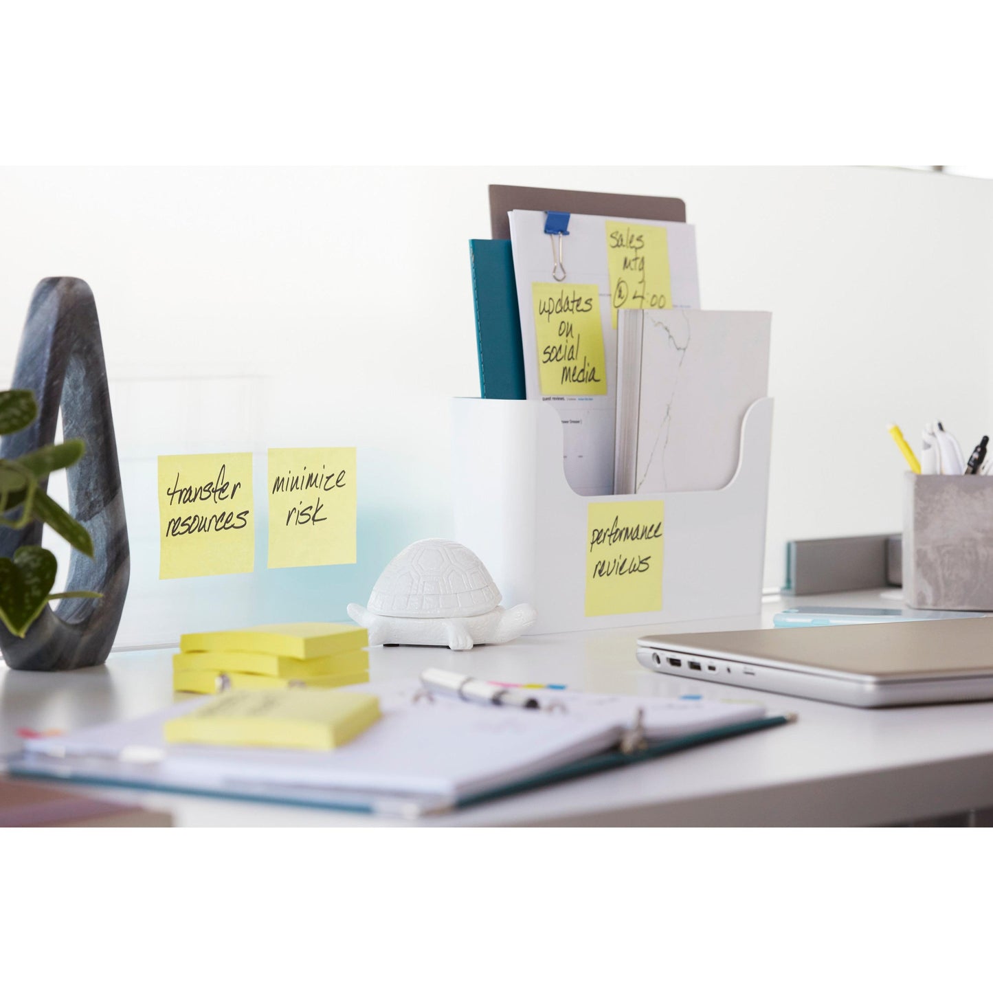 Egy rendezett irodai asztalon mappákkal, készletekkel, egy növénnyel és egy kis fehér teknősbéka figurával találhatóak a 3M Deutschland GmbH Post-it® Super Sticky Notes (sárga, 47,6 mm x 47,6 mm), ideális emlékeztetőknek, mint "Erőforrások áthelyezése" és "Kockázat minimalizálása".