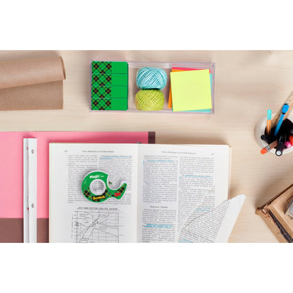 Ein Arbeitsbereich mit einem aufgeschlagenen Buch, Millimeterpapier, farbigen Garnkugeln, Haftnotizen, Stiften in einem Becher, Radiergummis, einer braunen Packpapierrolle und einer Rolle 3M Deutschland GmbH Scotch® Magic™ Unsichtbares Klebeband auf einem hellen Holztisch.