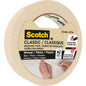 Egy tekercs 3M Scotch® Classic maszkolószalag, 24 mm x 50 m, 70% PEFC (SGSCH-COC-110078), ideális festési és felújítási munkákhoz fán, fémen, műanyagon. A magas tapadóerő akár két napig is tart. A képen egy kéz látható, amint a szalagot felhelyezi.