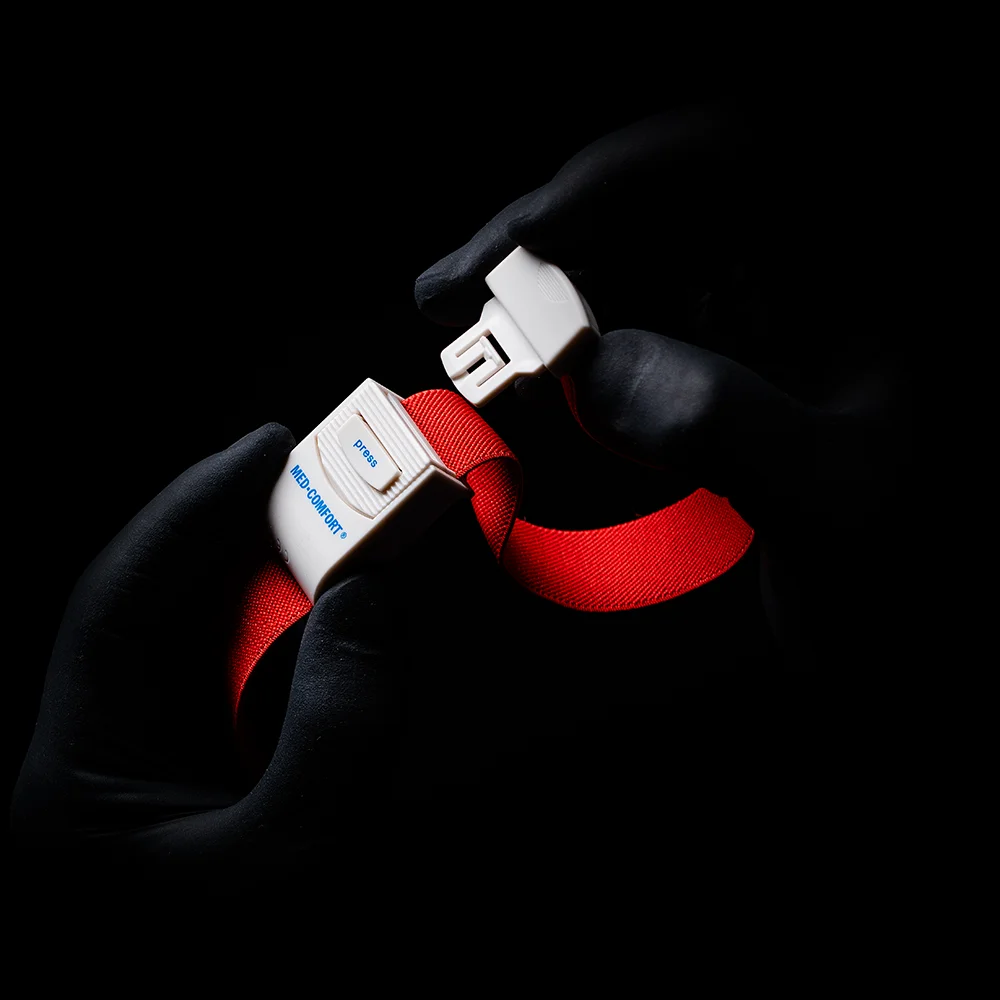 Kesztyűs kezek, amelyek egy AMPri MED-COMFORT vénaszorítóra emlékeztetnek, megfogják egy élénkpiros biztonsági öv csatját árnyékos háttér előtt. A csaton a „Blue” szó és a „B” betű látható.