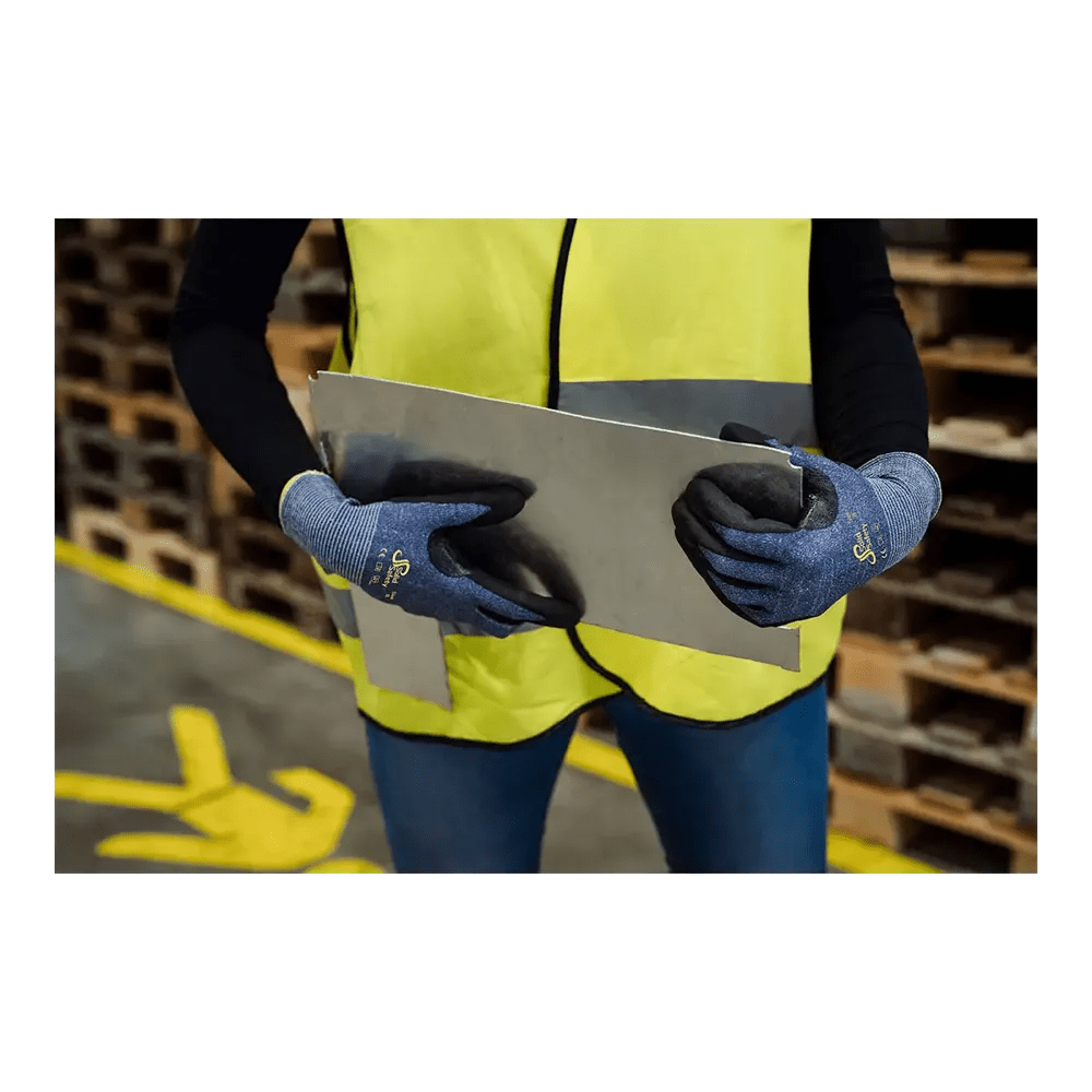 Egy személy neon sárga biztonsági mellényben és kék AMPri SolidSafety Cut vágásvédő kesztyűben S - XXL az AMPri Handelsgesellschaft mbH-tól óvatosan kezel egy nagy fémlemezt a raktárban. A háttérben fa raklapok vannak felhalmozva, a padlón lévő sárga jelzések biztonságos mozgásokat irányítanak és hangsúlyozzák a munkavédelmet.