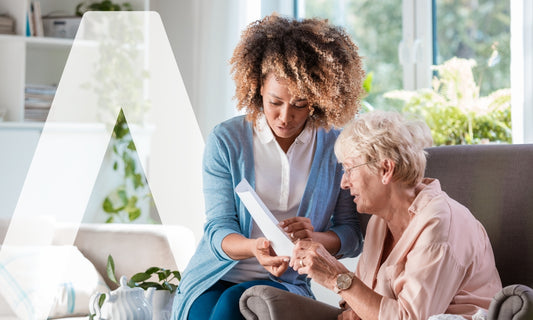Eine jüngere Frau zeigt einer älteren Frau, die auf einer Couch in einem hellen Wohnzimmer sitzt, ein Dokument oder ein Tablet. Sie scheinen gemeinsam Informationen zu besprechen.