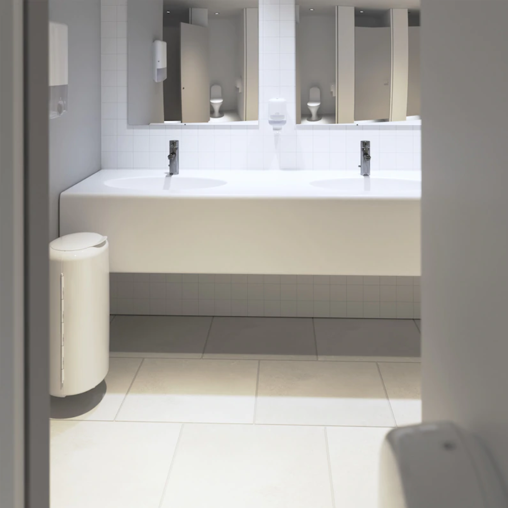 Egy modern nyilvános WC két fehér mosdóval rendelkezik, felettük téglalap alakú tükrökkel, amelyeket sima világosszürke csempék vesznek körül. Egy Tork 561000 Mini adagoló folyékony szappanhoz Elevation S2 az Essity Professional Hygiene Germany GmbH-tól a mosdók mellett van felszerelve, egy fehér szemetes társaságában, hogy tisztaságot biztosítson.