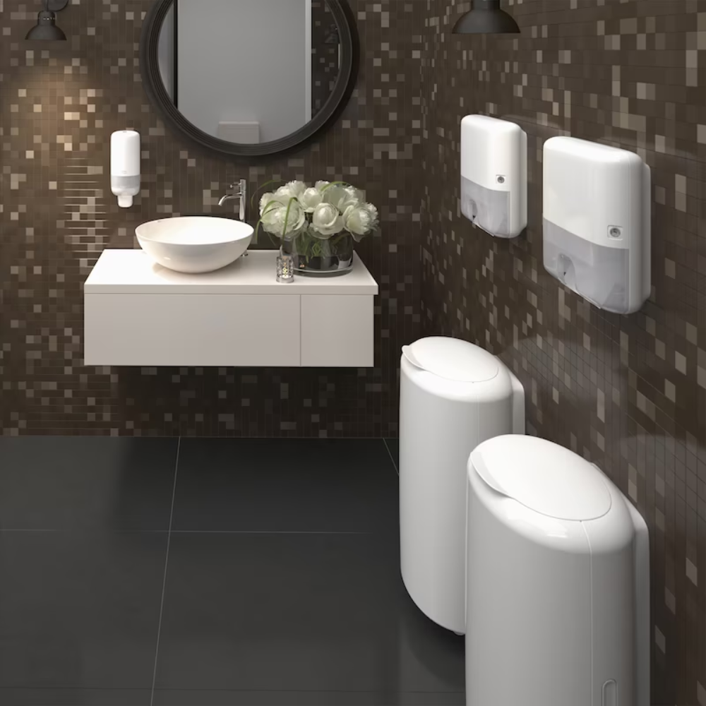Egy modern nyilvános WC barna mozaikcsempés fallal rendelkezik, amelyen egy kerek tükör, falra szerelt mosdókagyló fehér medencével, szappanadagoló, Tork Xpress® 552100 Mini adagoló Multifold kéztörlőkhöz Elevation H2 a TORK – Essity Professional Hygiene Germany GmbH-tól és két fehér hengeres szemetes található. Egy csokor fehér virág díszíti a mosdókagyló területét.