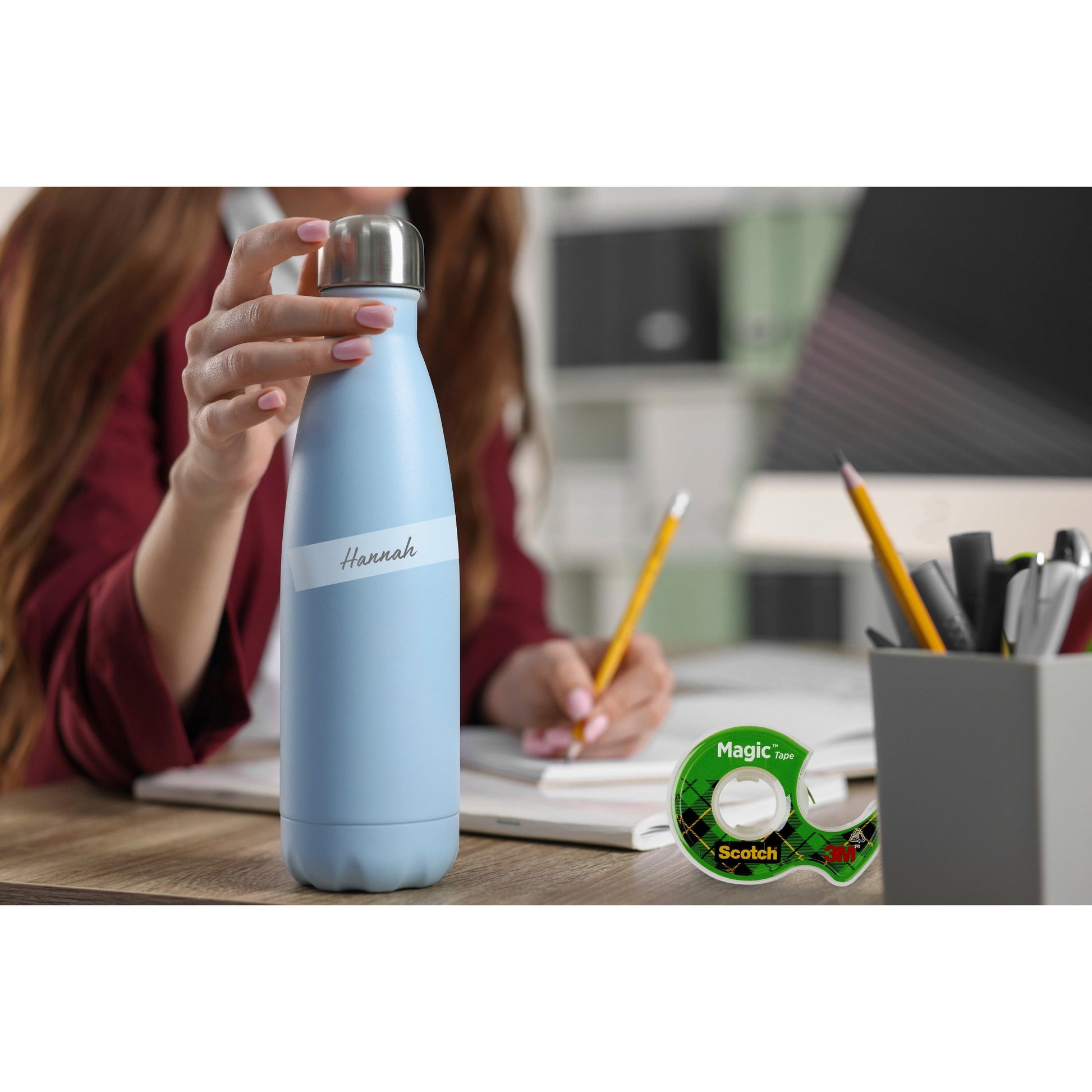 Eine Person hält eine hellblaue Wasserflasche mit der Aufschrift "Hannah" an einem Schreibtisch mit Büromaterial, einem Bleistift und einer Packung Scotch® Magic™ Unsichtbares Klebeband 8-1925R3 von 3M Deutschland GmbH (19 mm x 25 m, 3 Rollen) im Vordergrund.