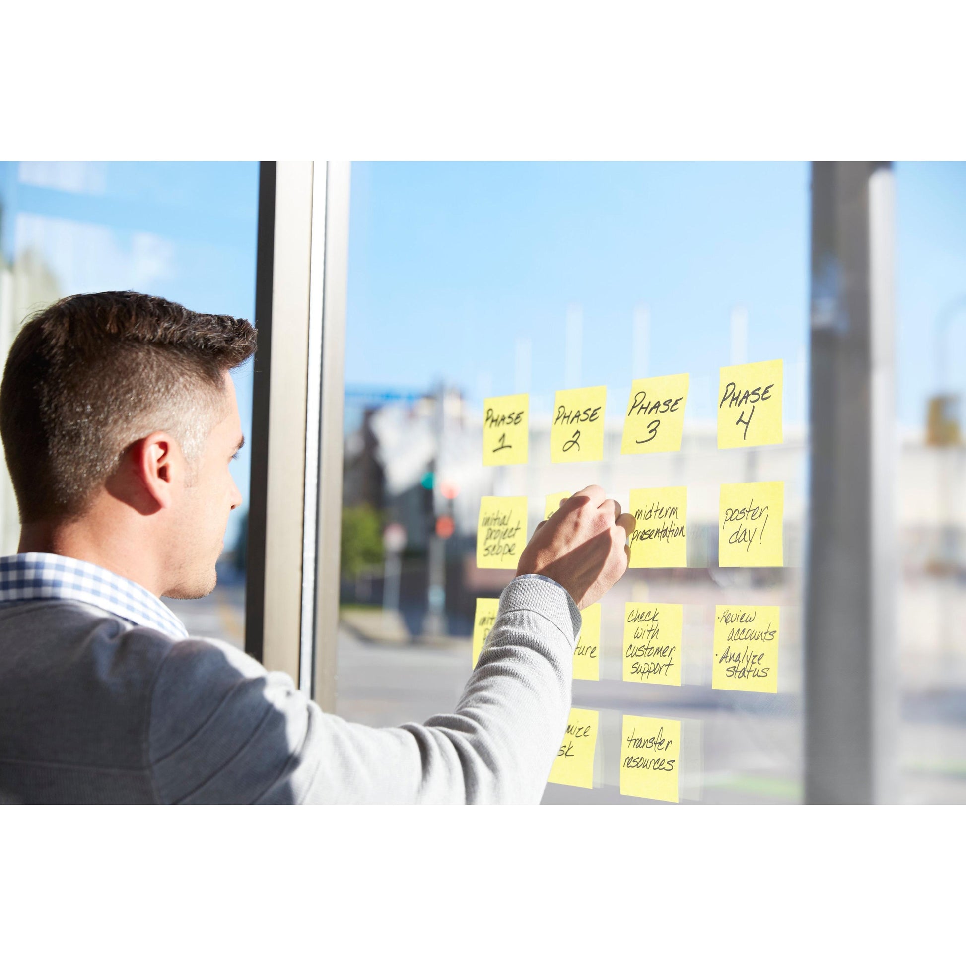 Eine Person organisiert Projektphasen und Aufgaben an einem Fenster mit Hilfe der Post-it® Super Sticky Z-Notes, Gelb, 76 x 76 mm, von 3M Deutschland GmbH, die in vier Spalten mit Phase 1-4 beschriftet sind. Sonnenlicht strömt herein und eine Straße ist draußen zu sehen.