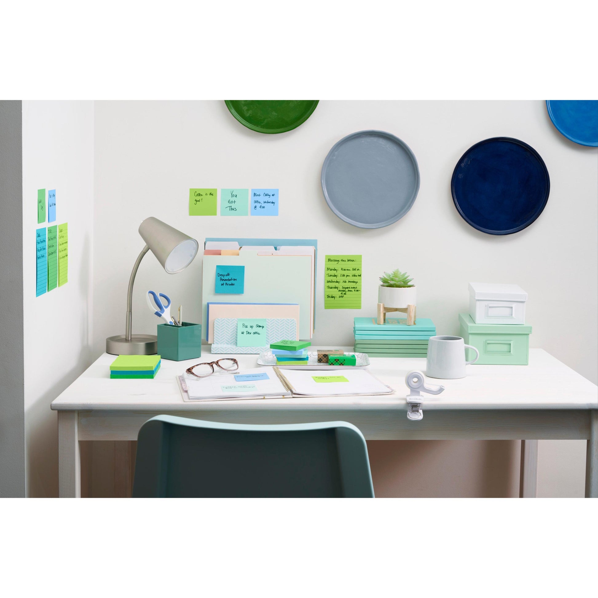 Ein aufgeräumter Schreibtisch mit organisiertem Büromaterial, Ordnern, einer Schreibtischlampe, einer Brille, einer Pflanze, Schachteln und Post-it® Super Sticky Notes (76 mm x 76 mm, 90 Blatt/Block) von 3M Deutschland GmbH. Darüber hängen bunte Teller und davor sitzt ein mintgrüner Stuhl.