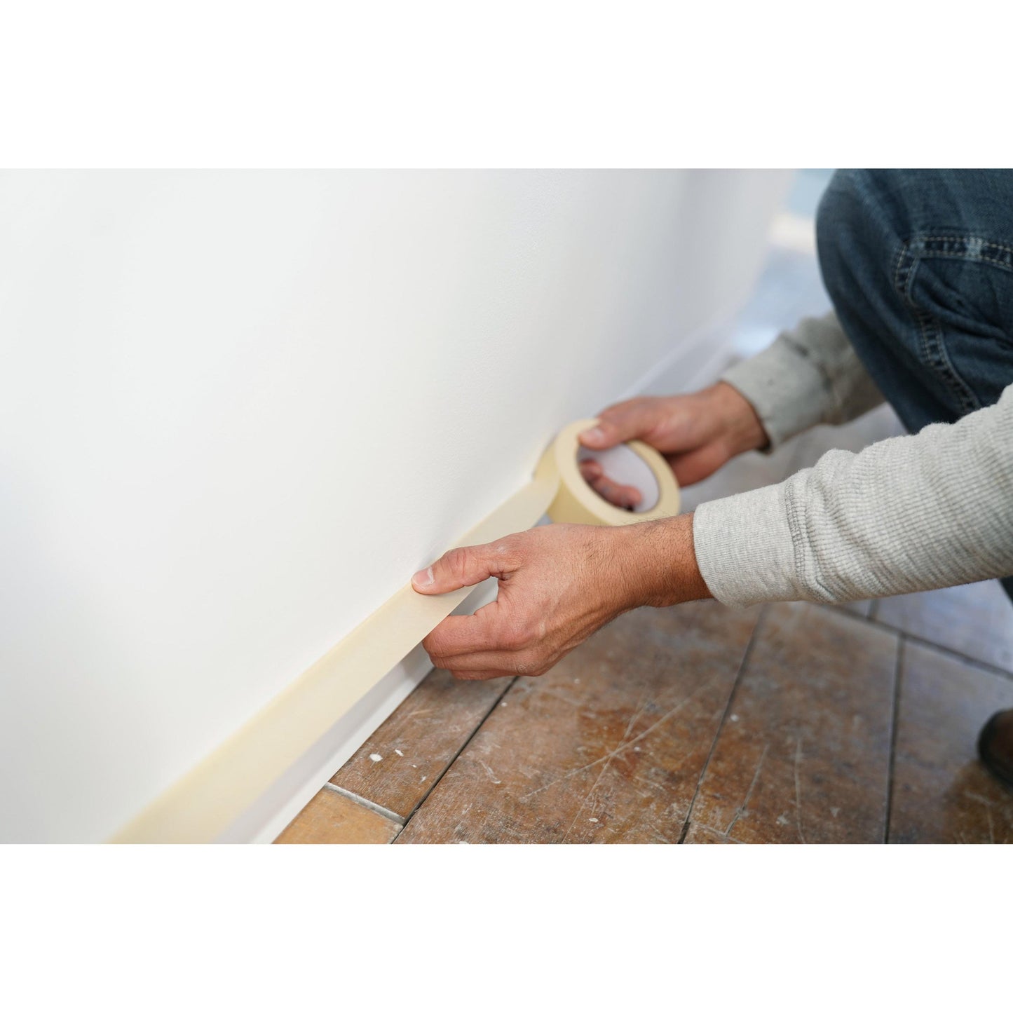 Egy személy a 3M Deutschland GmbH Scotch® Classic maszkolószalagját (24 mm x 50 m, 1 tekercs/csomag, 70% PEFC) használja, hogy festési és felújítási munkák során letakarja a fehér fal szegélyét egy csempézett padló felett.