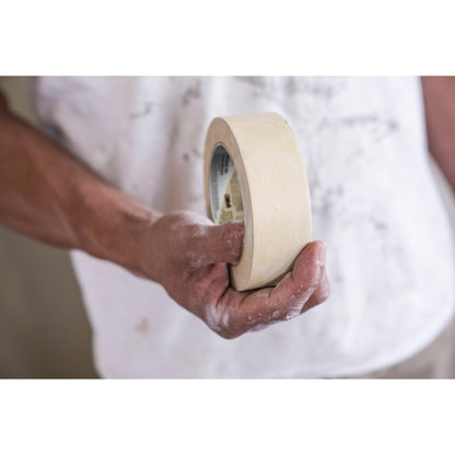 Egy személy tart egy tekercs Scotch® Classic maszkolószalagot (24 mm x 50 m, 70% PEFC, SGSCH-COC-110078) a 3M Deutschland GmbH-tól. Festékfoltos inget visel, és festék van a kezén, ami felújítási munkákra utal.