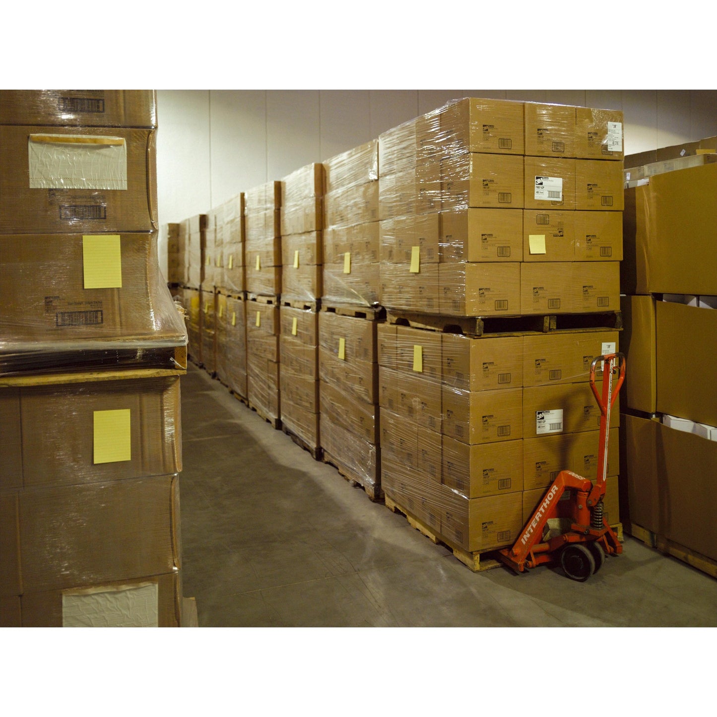 In einem Lagerhaus sind Stapel von Post-it® Super Sticky Notes im Großformat, Ultra Gelb (101 x 152 mm, 90 Blatt/Block, 6 Blöcke), von 3M Deutschland GmbH, auf Paletten verpackt. Ein roter Handhubwagen steht daneben.