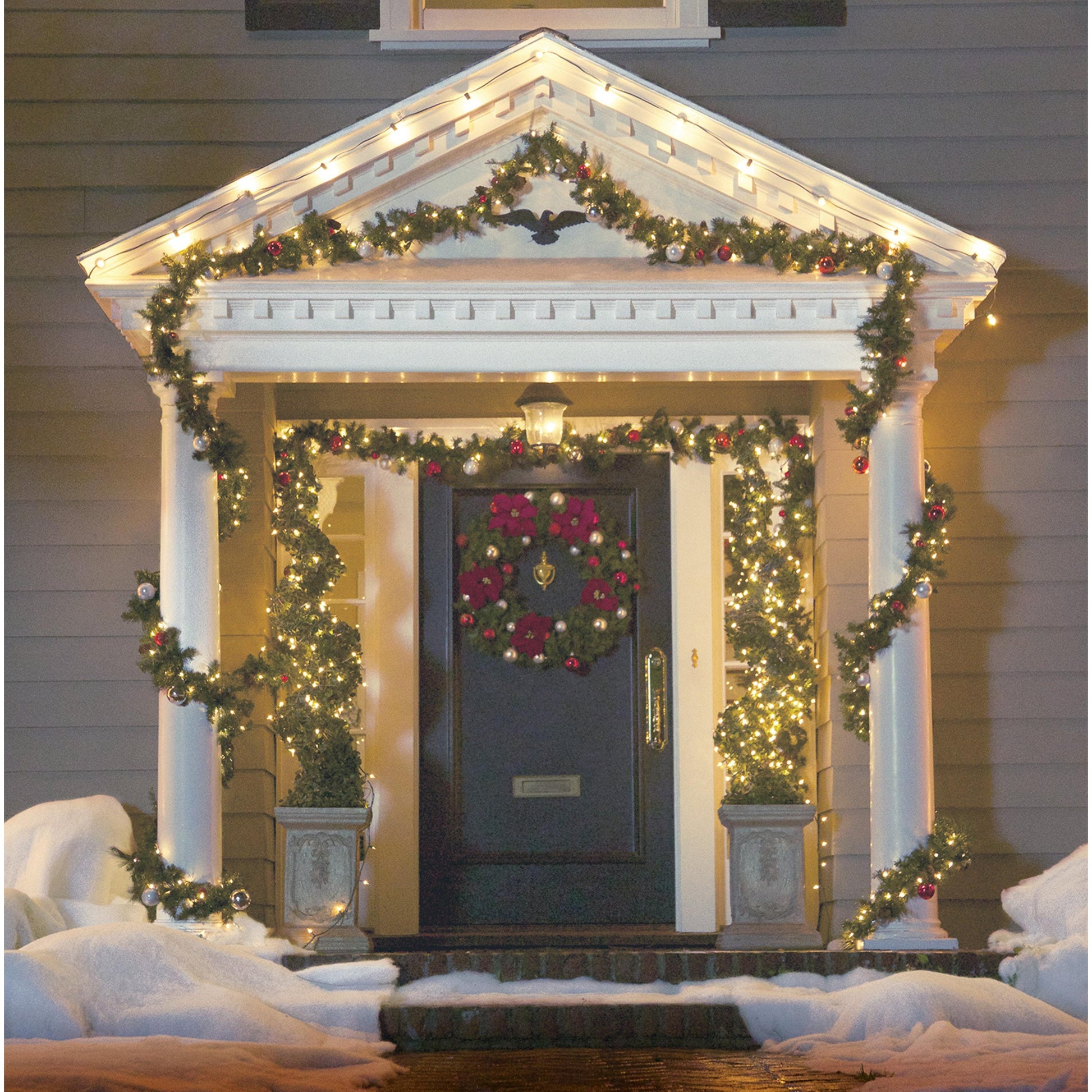 Eine weihnachtlich geschmückte Veranda mit Lichterketten, die mit Command™ Outdoor Lichter-Clips der 3M Deutschland GmbH sicher aufgehängt wurden. Der Boden ist mit Schnee bedeckt, und zwei Topfpflanzen flankieren den mit einem festlichen Kranz geschmückten Eingangsbereich.