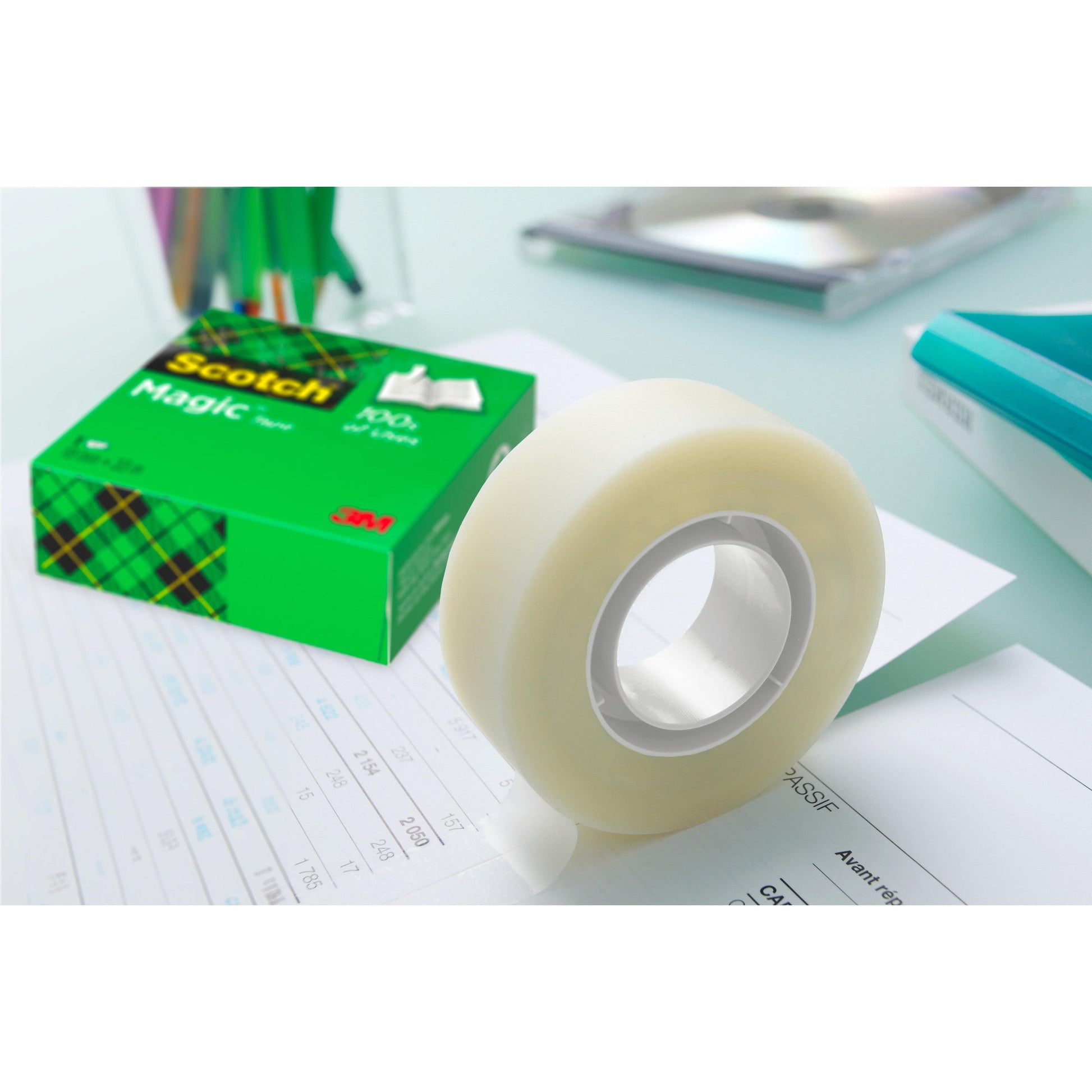 Eine Rolle Scotch® Magic™ Unsichtbares Klebeband von 3M liegt neben der grünen Vorteilspack-Packung (24 Rollen, 19 mm x 33 m) auf einem Schreibtisch mit sichtbaren Büroartikeln im Hintergrund.