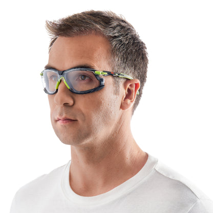 Ein Mann mit kurzen braunen Haaren trägt die 3M™ Solus™ Schutzbrille 1000 mit grünen und schwarzen Akzenten und klaren Gläsern von 3M Deutschland GmbH, leicht zur Seite gewandt in einem schlichten weißen T-Shirt vor weißem Hintergrund.